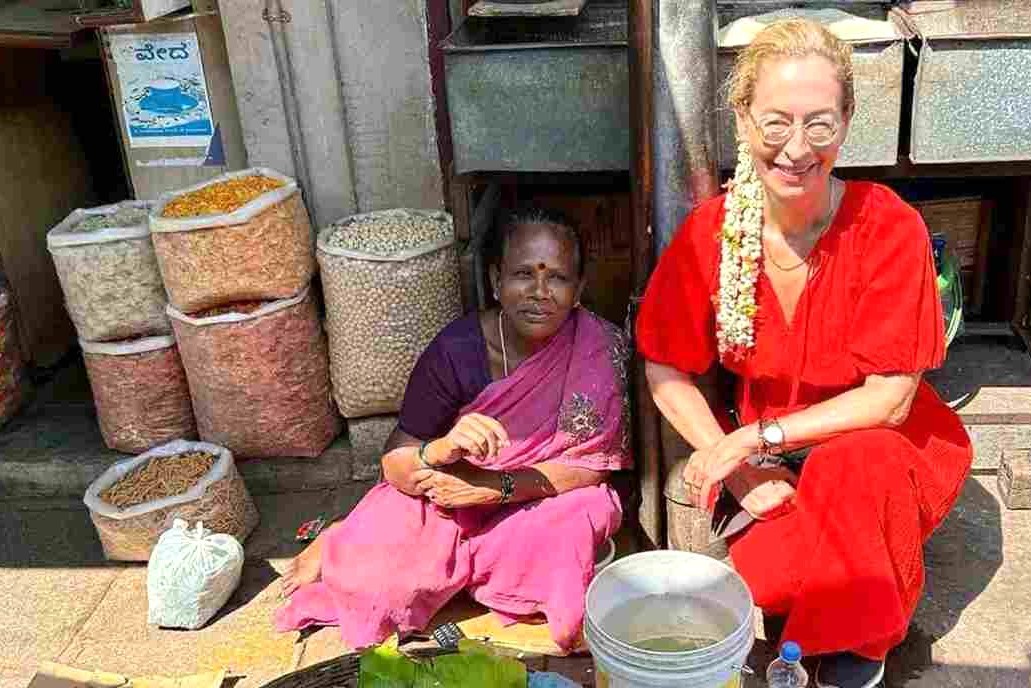 Local sari-clad vendor selling nuts and spices beside a smiling tourist at Devaraja Market in Mysore, Karnataka, India.