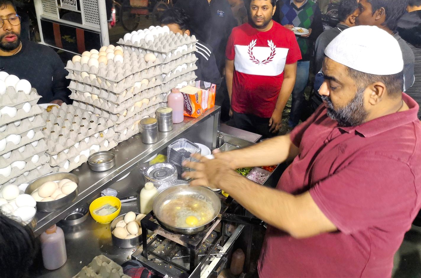 A bustling street food vendor at Agrasen Ki Baoli New Delhi, India skilfully prepares sizzling egg masala on a portable stove surrounded by eager diners, spices, trays, condiments and ambiance.