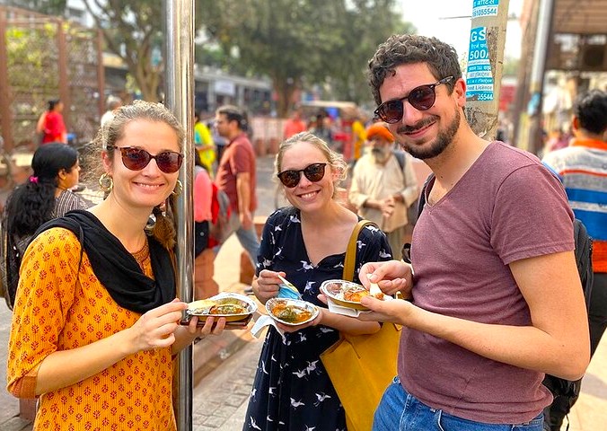 Three smiling young travelers savoring authentic Indian street food at the historic Agrasen Ki Baoli stepwell in New Delhi, India, capturing vibrant local culture, heritage architecture and lively marketplace atmosphere.
