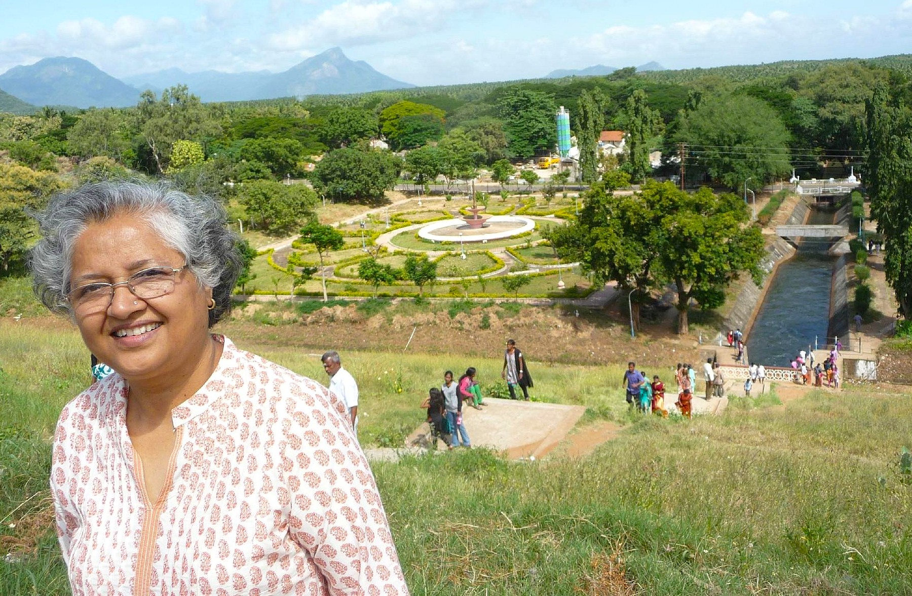 Scenic view of Palani Aliyar Dam reservoir in Tamil Nadu, India, with lush hills, mountain backdrop, winding canal footbridge and visitors strolling along grassy embankments. Scenic view of Palani Aliyar Dam reservoir in Tamil Nadu, India, with lush hills, mountain backdrop, winding canal footbridge and visitors strolling along grassy embankments.