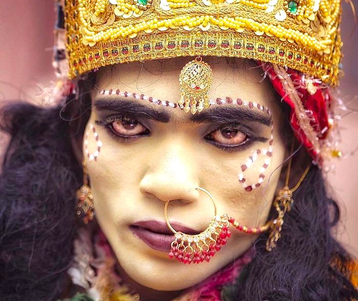 A striking portrait of a traditional divine performer at Ambrai Ghat, Udaipur, Rajasthan, India, showcasing ornate jewelry, vibrant makeup, cultural symbolism, expressive gaze, and intricate headgear against serene lakefront ambiance. A striking portrait of a traditional divine performer at Ambrai Ghat, Udaipur, Rajasthan, India, showcasing ornate jewelry, vibrant makeup, cultural symbolism, expressive gaze, and intricate headgear against serene lakefront ambiance.