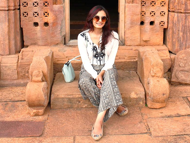 Here is an SEO-optimized sentence based on your request:  A fashionable female tourist explores the ancient temple complex in Badami, Karnataka, India, sitting on the carved stone steps. This historic location, near the famous Pattadakal UNESCO World Heritage site, is a prime example of India's rich architectural heritage and a top destination for travelers.