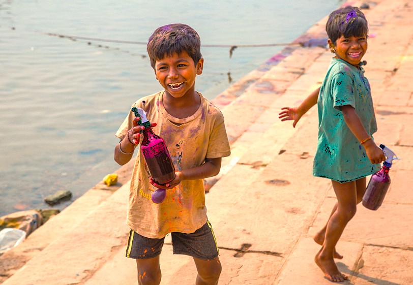 “Two children play and spray water joyfully by the riverbank in Varanasi, Uttar Pradesh, India, near Sarnath Temple Buddha and Bharat Kala Bhavan Museum, capturing youthful exuberance and festival fun.”