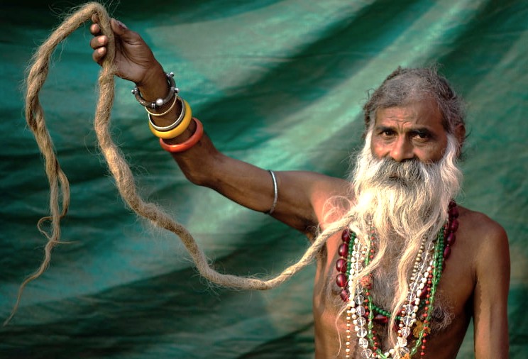 An elderly Rajasthani man with a long white beard and braided hair holds strands adorned with beads and bangles during the Bikaner Camel Safari in India’s Thar Desert of Rajasthan.