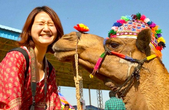 A joyful tourist in vibrant traditional Rajasthani attire shares a playful moment with a colorfully adorned camel at the Bikaner Camel Festival in the sunlit Thar Desert of Rajasthan, India.