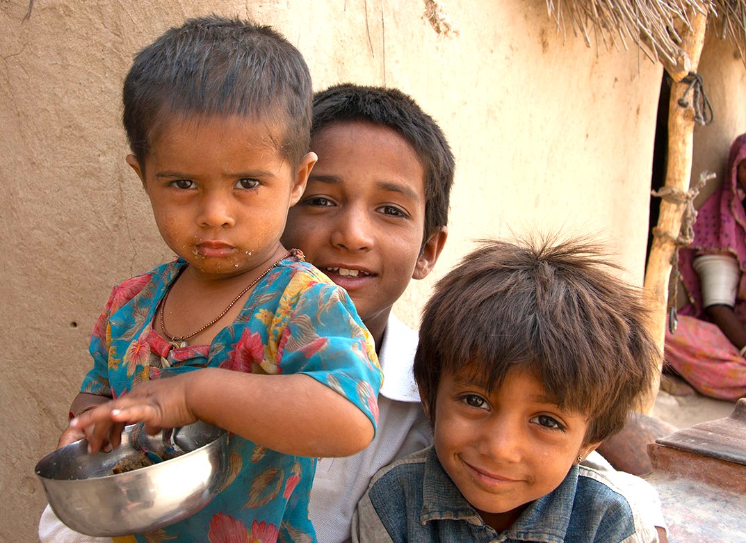 Three smiling rural children enjoying a meal in a village in Rajasthan, India, near the vast Thar Desert, evoking community spirit and cultural heritage on a Bikaner Camel Safari experience.