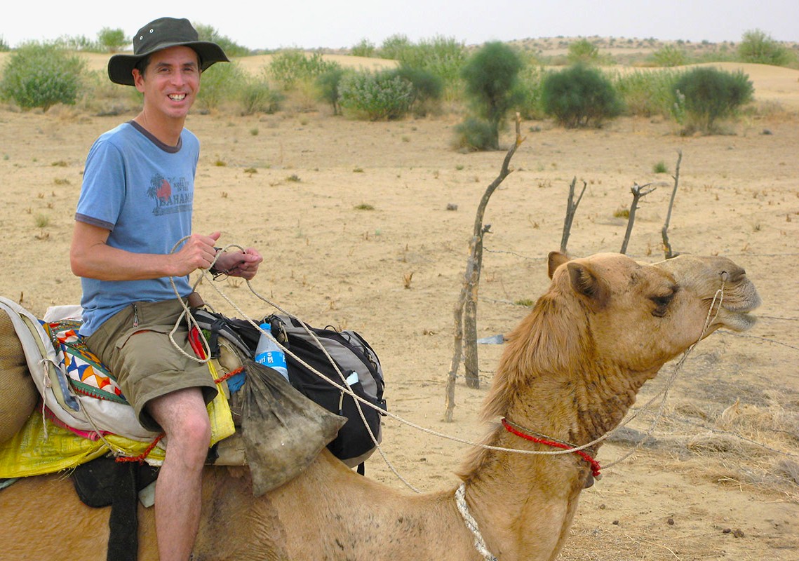 A smiling traveler on a traditional camel in the arid Thar Desert of Rajasthan, India, enjoying an authentic Bikaner Camel Safari adventure amidst golden dunes and desert landscapes beneath skies.