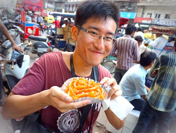 Smiling tourist holding crispy orange jalebi snack in bustling India-Chandi Chowk New Delhi street market, surrounded by colorful stalls, busy vendors, local shoppers, motorbikes, traditional heritage architecture, vibrant culinary culture. Smiling tourist holding crispy orange jalebi snack in bustling India-Chandi Chowk New Delhi street market, surrounded by colorful stalls, busy vendors, local shoppers, motorbikes, traditional heritage architecture, vibrant culinary culture.