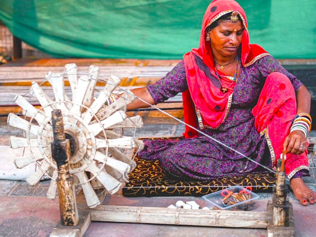 Local Rajasthani artisan weaving cotton threads on a traditional spinning wheel outside Jaipur’s City Palace, Rajasthan, India, UNESCO heritage site, showcasing vibrant cultural craftsmanship, colorful attire, and historic market traditions.