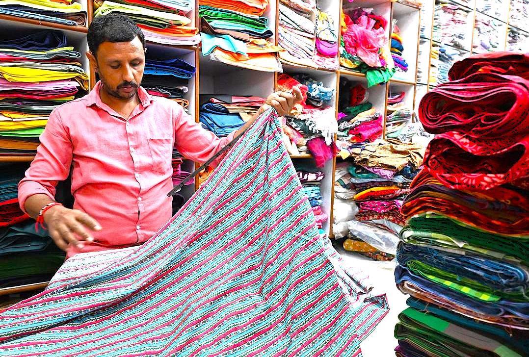 Colorful fabric shop in Coimbatore Tamil Nadu, India, with vendor inspecting vibrant cotton and silk textiles on neatly organized shelves, highlighting local textile craftsmanship patterns. Colorful fabric shop in Coimbatore Tamil Nadu, India, with vendor inspecting vibrant cotton and silk textiles on neatly organized shelves, highlighting local textile craftsmanship patterns.