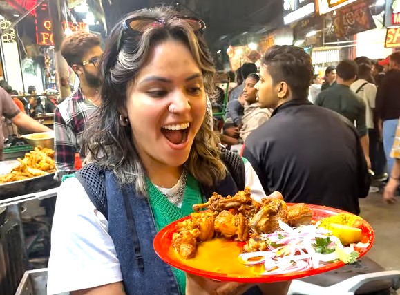 Enthusiastic young woman savoring spicy street food at India-Chandi Chowk New Delhi night market, vibrant fried chicken and onion salad on red plate, bustling vendors, neon signage, urban culinary adventure. Enthusiastic young woman savoring spicy street food at India-Chandi Chowk New Delhi night market, vibrant fried chicken and onion salad on red plate, bustling vendors, neon signage, urban culinary adventure.