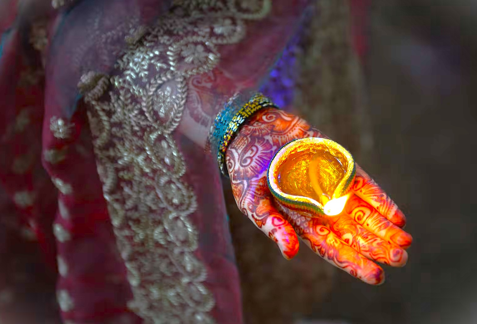 “Close-up of henna-adorned hand holding glowing diya oil lamp during Diwali festival at Ambrai Ghat waterfront in Udaipur, Rajasthan, India, capturing ornate traditional attire, cultural ritual, warm, luminous, serene atmosphere.”  “Close-up of henna-adorned hand holding glowing diya oil lamp during Diwali festival at Ambrai Ghat waterfront in Udaipur, Rajasthan, India, capturing ornate traditional attire, cultural ritual, warm, luminous, serene atmosphere.”