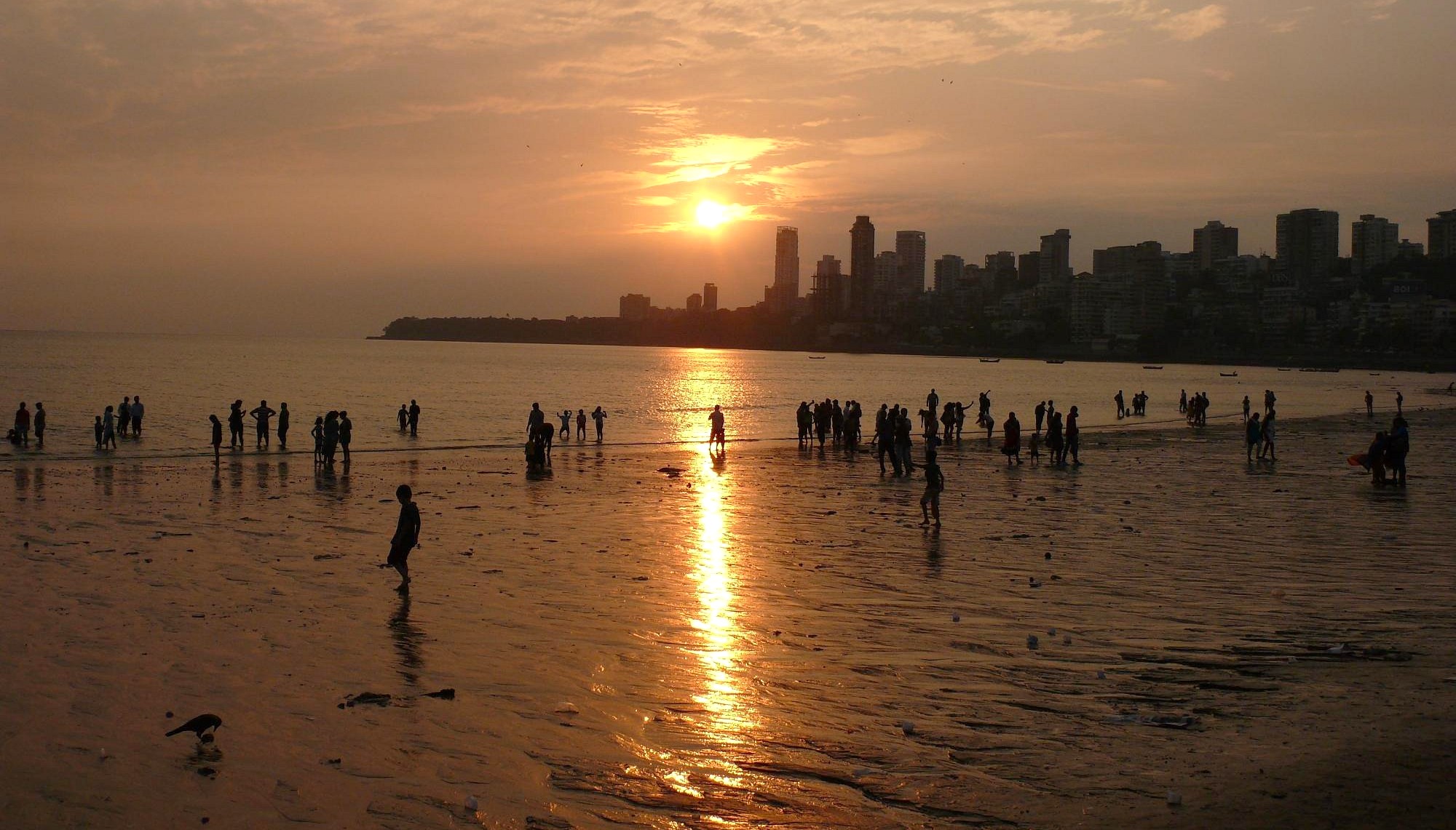 Vibrant sunset at Chowpatty Beach in Mumbai, India showcases tranquil waves, reflective wet sand, silhouetted visitors and the iconic Mahapradesh skyline, inviting cultural exploration and seaside relaxation under golden skies.
