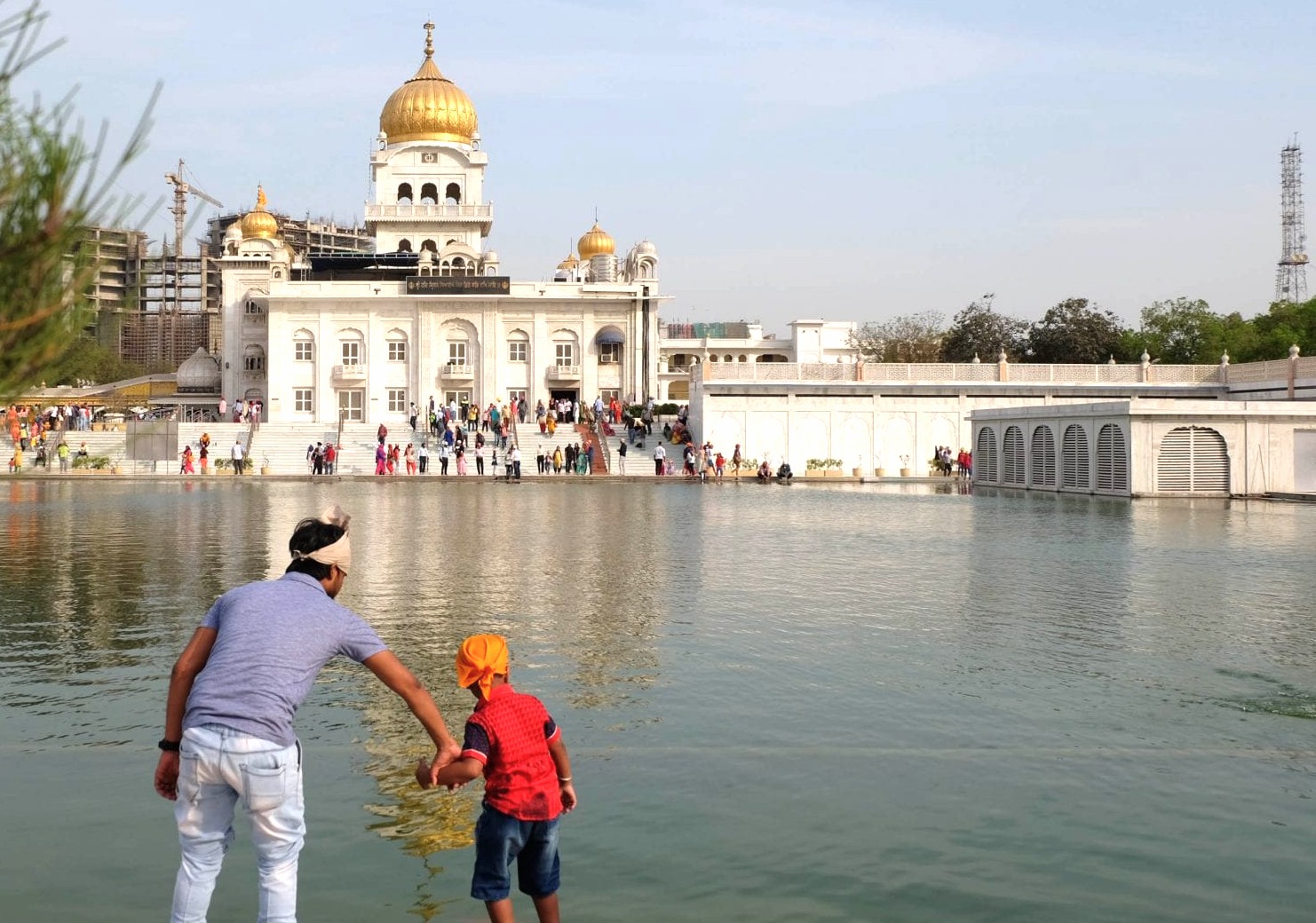 Father guiding child to toss offering into serene sarovar waters at Gurudwara Bangla Sahib Sikh Temple in New Delhi, India, reflecting spiritual solace and community devotion before white marble shrine. Father guiding child to toss offering into serene sarovar waters at Gurudwara Bangla Sahib Sikh Temple in New Delhi, India, reflecting spiritual solace and community devotion before white marble shrine.