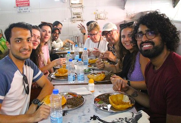  “International diverse visitors at New Delhi’s Hanuman Mandir langar hall enjoying authentic traditional North Indian thali with puri, chole, lassi and communal dining atmosphere near sacred Hanuman shrine in India.”