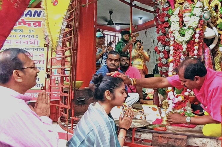 Devotees engaged in a Hanuman Mandir blessing ceremony in New Delhi, India, where a priest applies red tika to a young woman’s forehead amid floral decorations, traditional attire, devotional offerings.