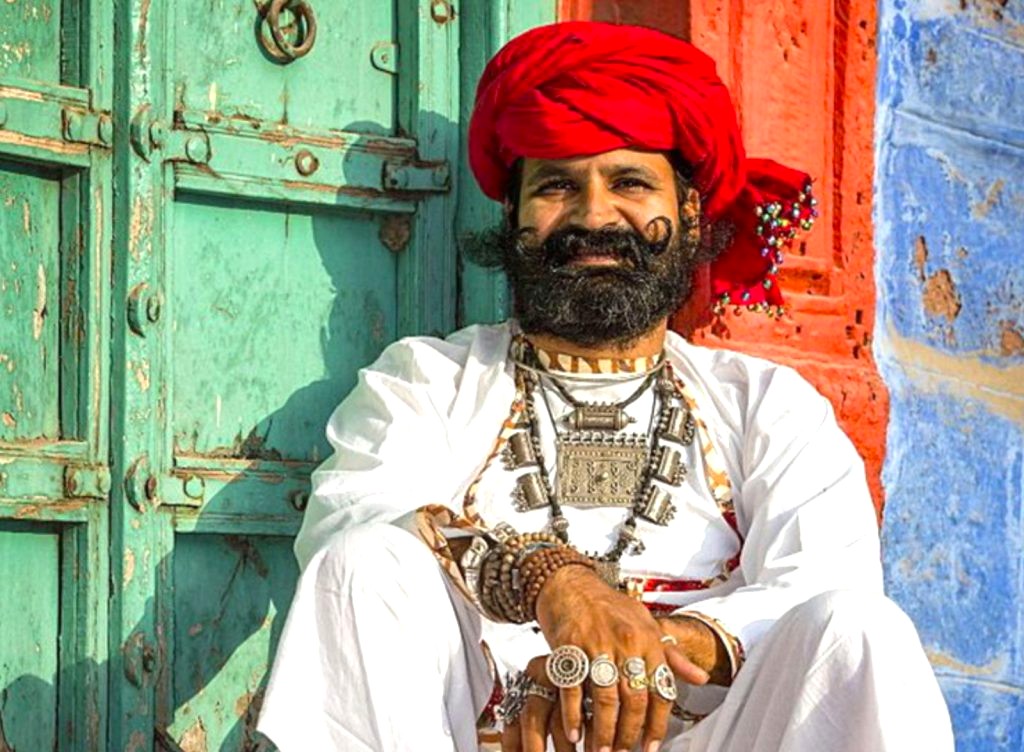 Rajasthani man wearing vibrant red pagri and traditional white attire sits against a colorful backdrop in Jaipur, Rajasthan, India near the UNESCO-listed Hawa Mahal–Palace of the Winds, showcasing cultural heritage.