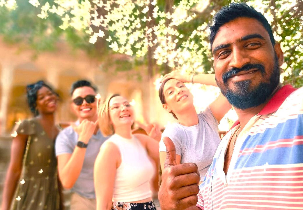 Five diverse friends smiling under blooming trees at Jaipur’s City Palace in Rajasthan, India, capture a joyful UNESCO heritage site visit, highlighting vibrant culture, historic architecture, and unforgettable travel memories.