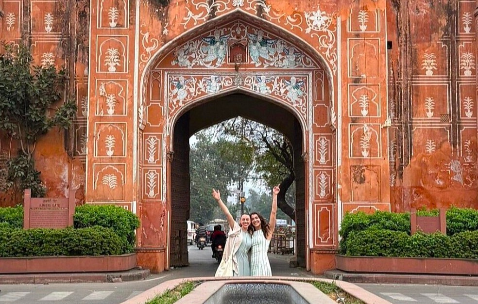 Two joyful women pose beneath an intricately carved sandstone gate in Jaipur near Hawa Mahal – Palace of the Winds, Rajasthan, India, celebrating cultural heritage at an iconic UNESCO landmark.