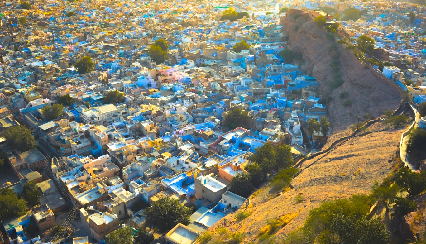 Aerial view of Jodhpur, the Blue City in Rajasthan, India, nestled on the edge of the Thar Desert, showcasing vibrant blue rooftops against arid rocky hills under golden evening light. Aerial view of Jodhpur, the Blue City in Rajasthan, India, nestled on the edge of the Thar Desert, showcasing vibrant blue rooftops against arid rocky hills under golden evening light.