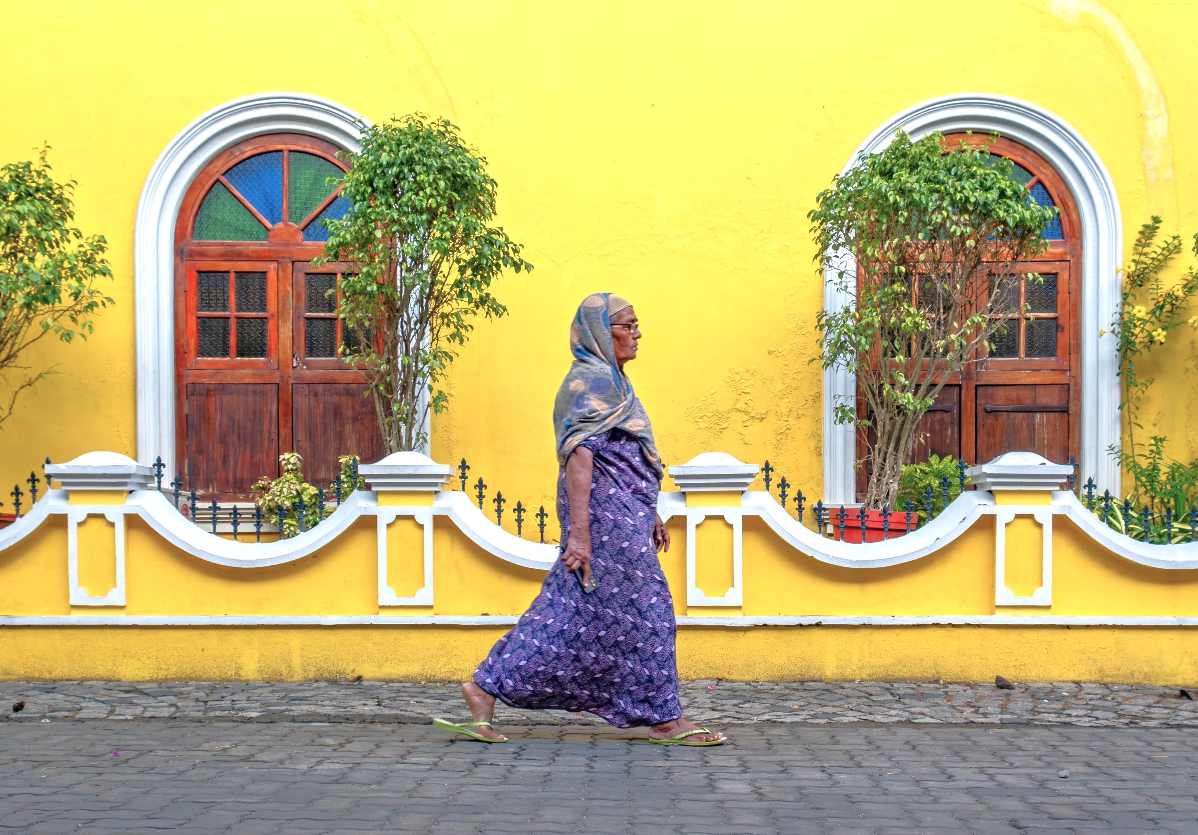 An elderly woman in traditional attire from South India strides past a vibrant yellow colonial facade in Kochi, Kerala, showcasing street life, cultural heritage, colorful architecture, routine and local authenticity.