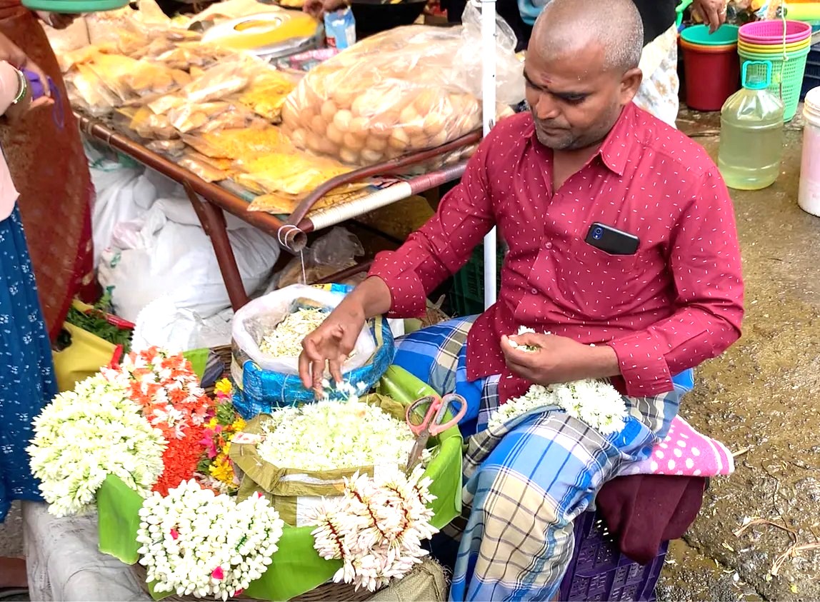 A vendor at the Kodaikanal Sunday Market in Tamil Nadu demonstrates his relationship with tradition by making fresh jasmine garlands, a cultural highlight for tourists in India exploring Madurai, Meenakshi Temple, and Munnar near the Unesco Western Ghats.