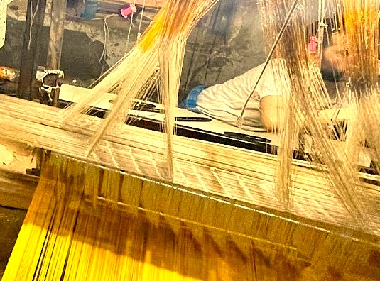 A weaver at Mehrotra-silks in Varanasi, India, demonstrates the intimate relationship between artisan and loom, crafting fine silk near the historic Sarnath UNESCO site in a time-honored tradition.