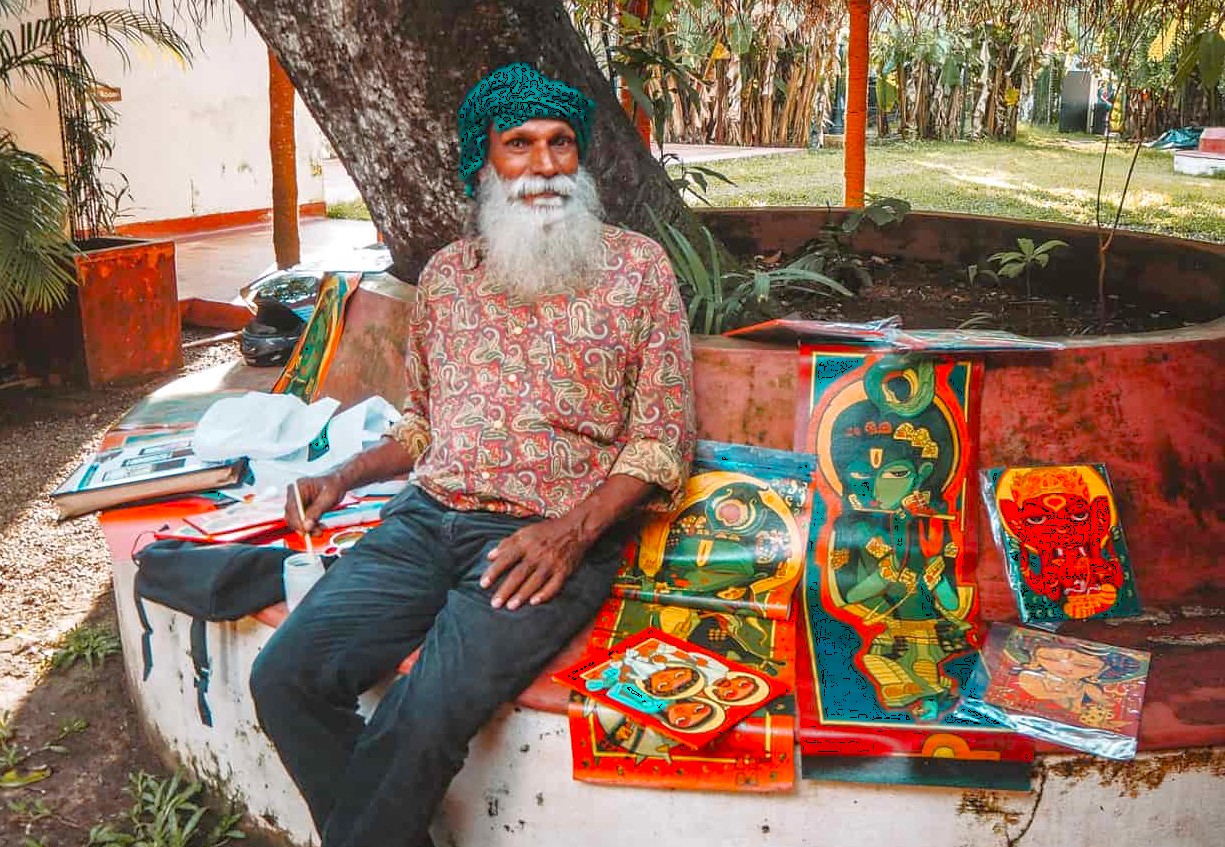 Elderly artist painting colorful vibrant Hindu mythological posters under a banyan tree at Munnar Kerala stall, Southern India, showcasing traditional craftsmanship, spiritual culture and heritage.