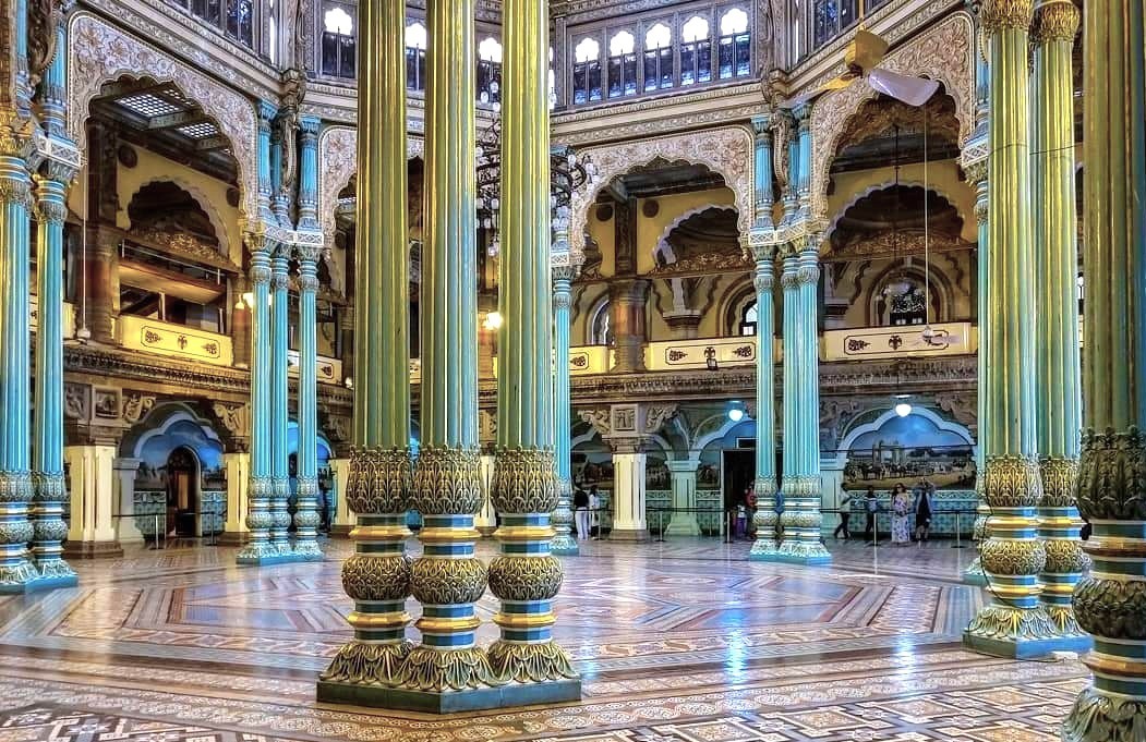 Magnificent ornate hall of Mysore Palace in Karnataka, India features towering green-gold columns, intricate arches, mosaic floors, stained-glass skylights, royal murals, sumptuous heritage architecture interior.