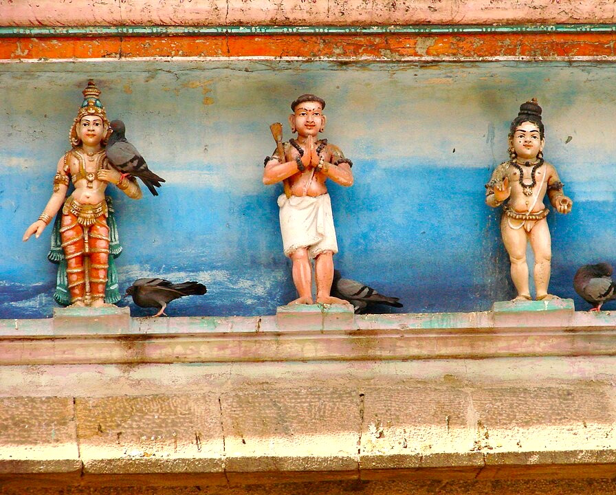 Three weathered painted deity sculptures on a rustic blue ledge at the Nadukkattu Gopuram of Meenakshi Temple in Madurai, Tamil Nadu, South India, captured alongside urban pigeons highlighting artistic heritage. Three weathered painted deity sculptures on a rustic blue ledge at the Nadukkattu Gopuram of Meenakshi Temple in Madurai, Tamil Nadu, South India, captured alongside urban pigeons highlighting artistic heritage.