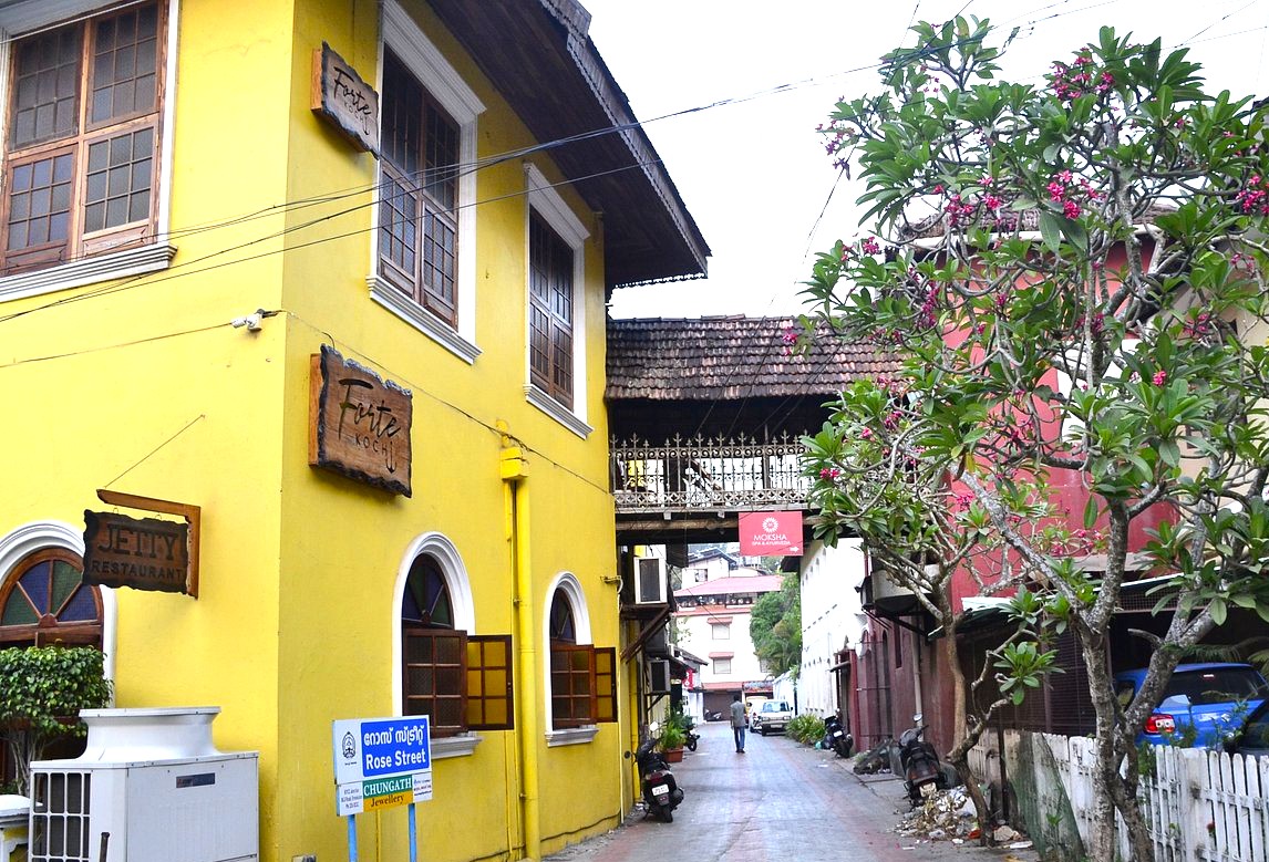 Vibrant street scene on Princess Street Kochi in Kerala, South India, featuring colonial yellow building with Jetty Restaurant and Forte Kochi signage, flowering tree, parked scooters, and quaint pedestrian atmosphere. Vibrant street scene on Princess Street Kochi in Kerala, South India, featuring colonial yellow building with Jetty Restaurant and Forte Kochi signage, flowering tree, parked scooters, and quaint pedestrian atmosphere.