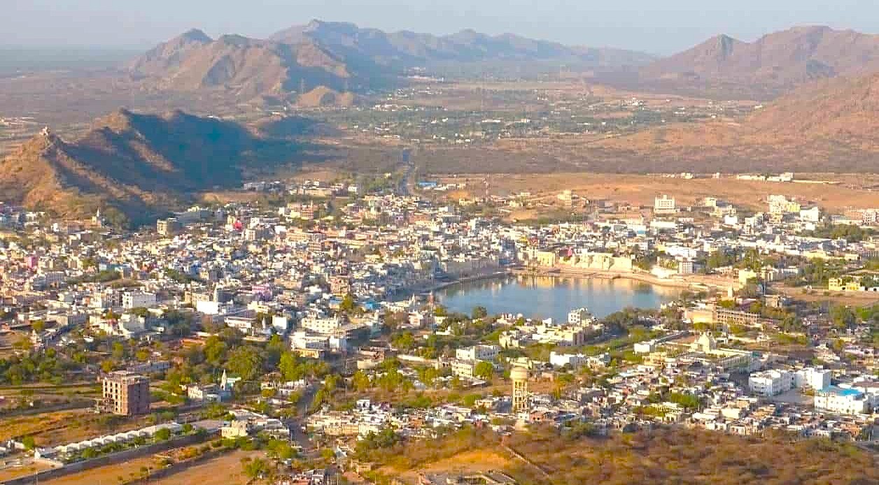 Aerial panorama of Pushkar in Rajasthan, India, nestled between Jaipur and Agra en route to the Taj Mahal, showcases a desert cityscape with temples, a sacred lake, and Aravalli foothills.