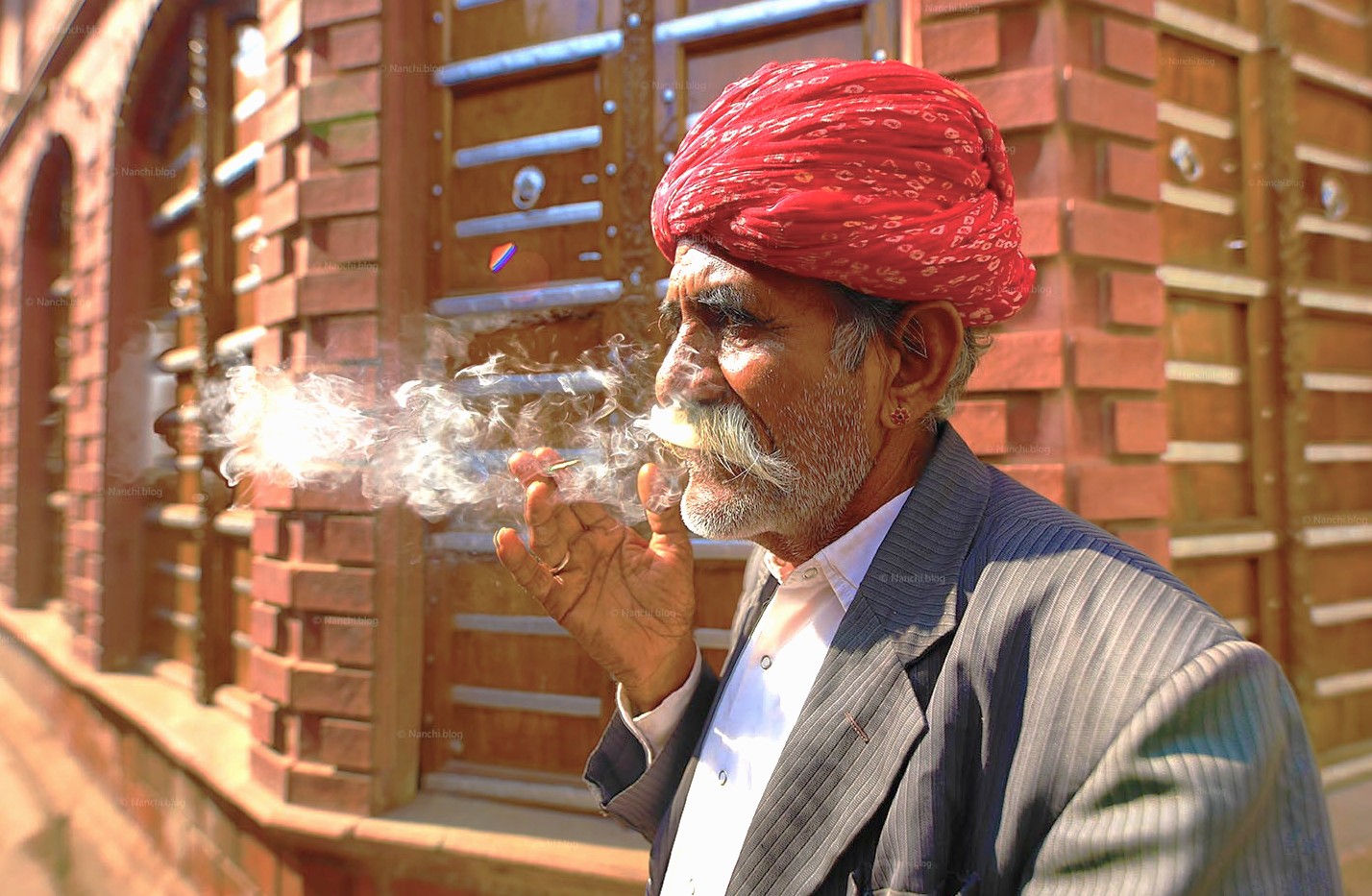 Elderly local resident smoking a traditional cigarette against vibrant ornate red Rampura Havelis facade in Bikaner, showcasing authentic street life and heritage architecture in Rajasthan’s Thar Desert region of India. Elderly local resident smoking a traditional cigarette against vibrant ornate red Rampura Havelis facade in Bikaner, showcasing authentic street life and heritage architecture in Rajasthan’s Thar Desert region of India.