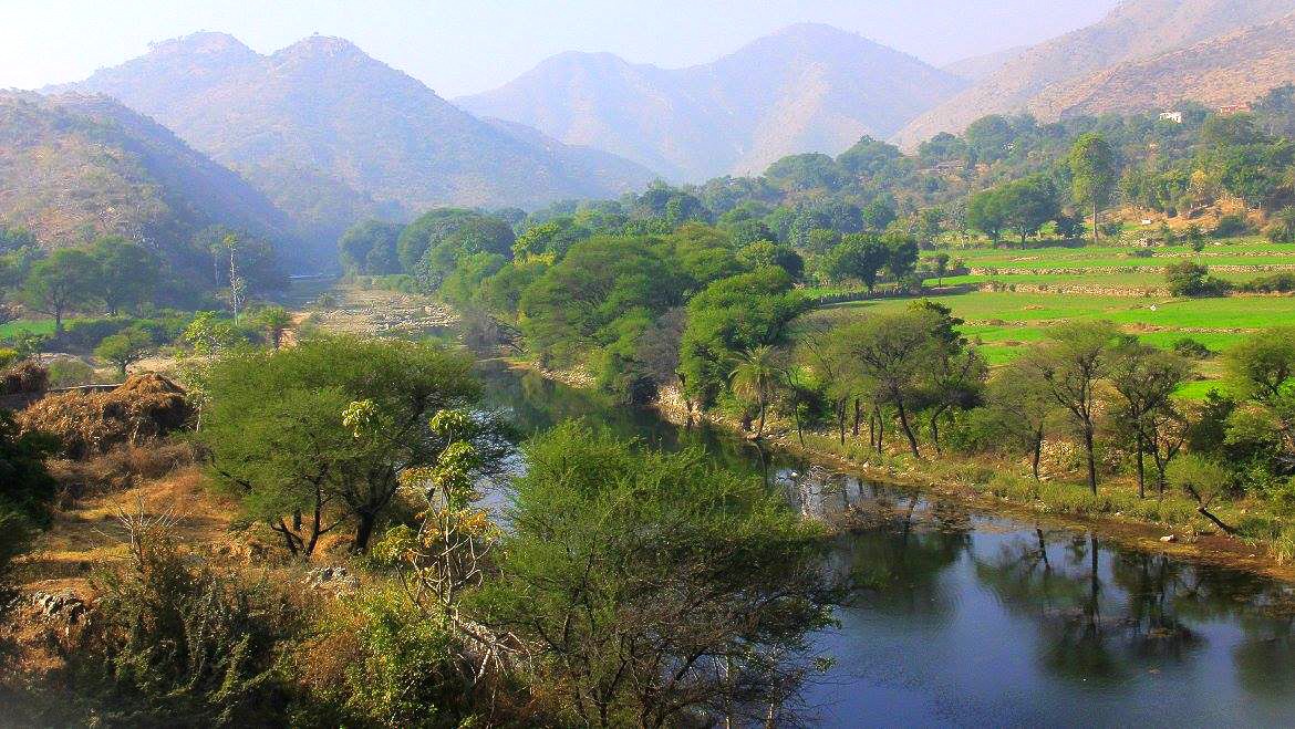 A scenic river valley winding through the Aravali Hills near Jodhpur, Rajasthan, India, overlooks terraced fields and distant Ranakpur Jain Temple, highlighting lush greenery, rugged mountains, and serene rural landscapes. A scenic river valley winding through the Aravali Hills near Jodhpur, Rajasthan, India, overlooks terraced fields and distant Ranakpur Jain Temple, highlighting lush greenery, rugged mountains, and serene rural landscapes.