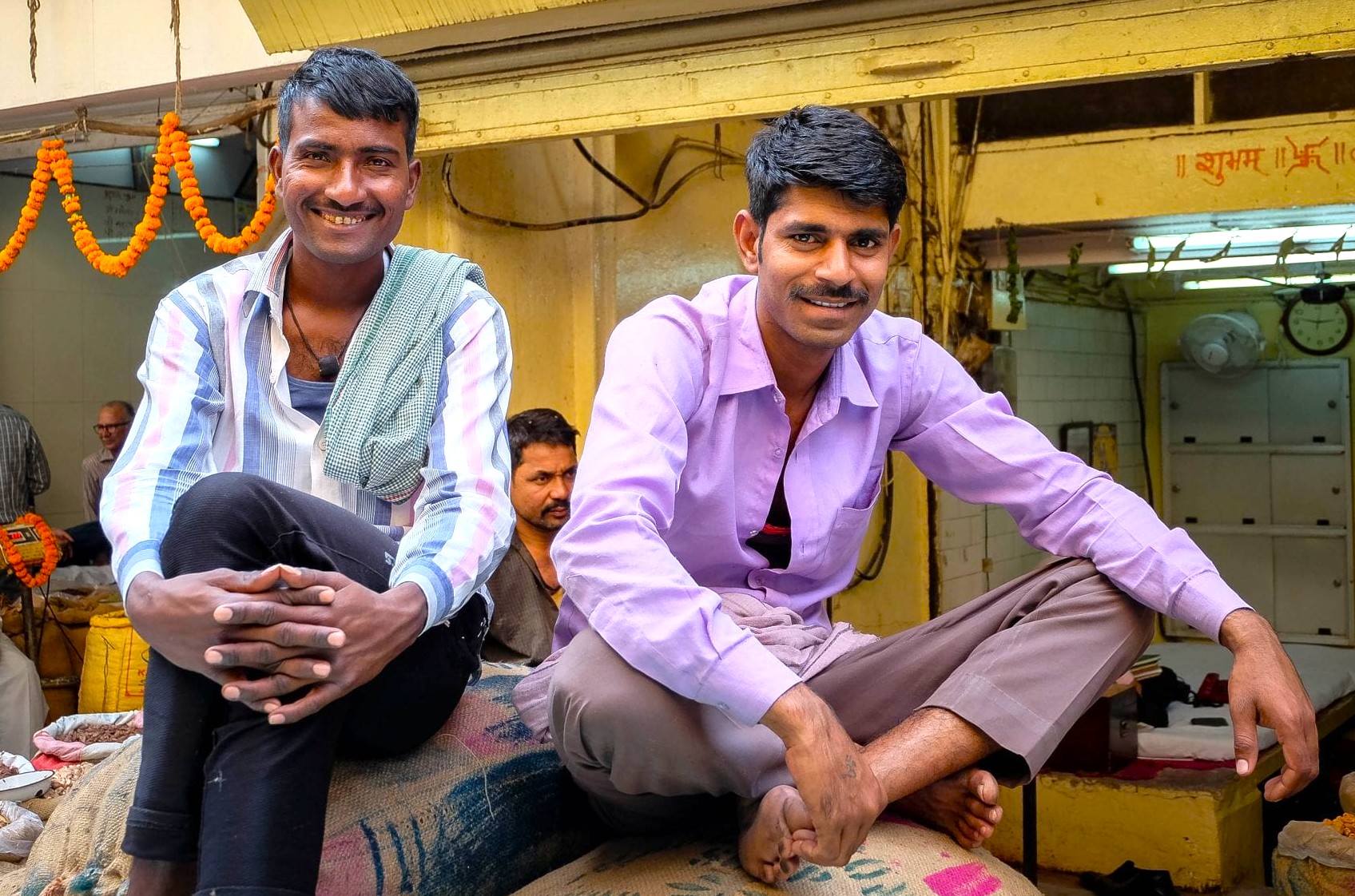 Two smiling street vendors sit on sacks in a vibrant marketplace near the Red Fort in Delhi, India, showcasing local culture, traditional attire, bustling bazaar, commerce, tourism, Chandni Chowk experience. Two smiling street vendors sit on sacks in a vibrant marketplace near the Red Fort in Delhi, India, showcasing local culture, traditional attire, bustling bazaar, commerce, tourism, Chandni Chowk experience.