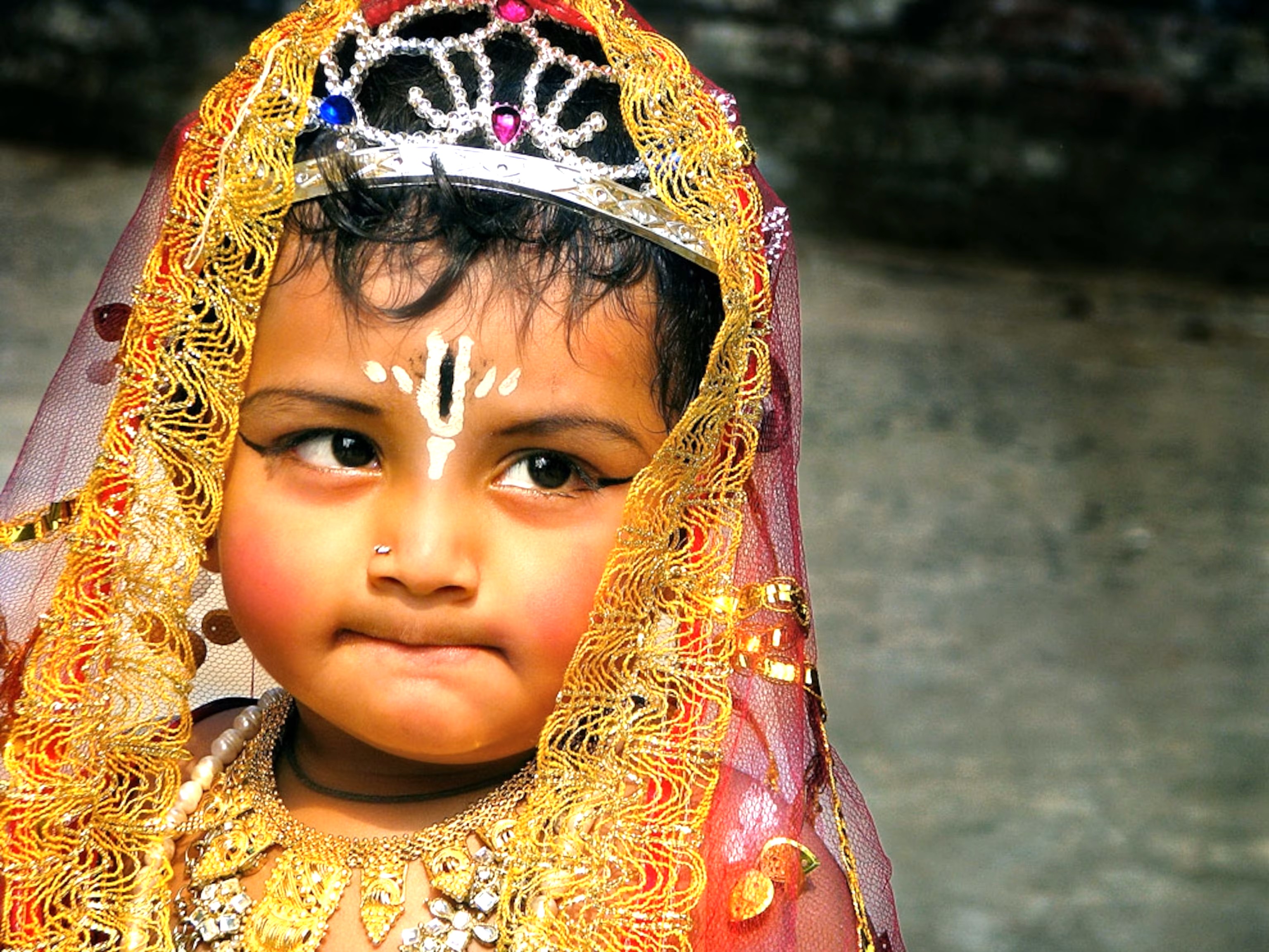 A young child in ornate ceremonial attire and facial markings stands before the Royal Cenotaphs in Bikaner, Rajasthan, India, against the arid Thar Desert backdrop, capturing vibrant timeless cultural heritage. A young child in ornate ceremonial attire and facial markings stands before the Royal Cenotaphs in Bikaner, Rajasthan, India, against the arid Thar Desert backdrop, capturing vibrant timeless cultural heritage.