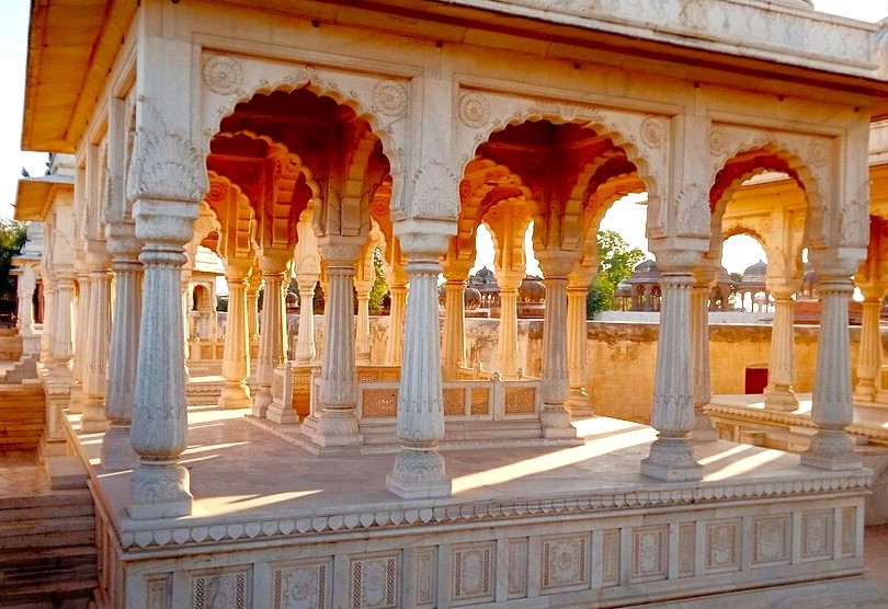 Intricately carved white marble Royal Cenotaphs in Bikaner, Rajasthan, India, stand amid the arid Thar Desert landscape, showcasing Mughal-inspired arches, ornate columns, and historic Rajasthani architectural splendor, illuminated by sunlight. Intricately carved white marble Royal Cenotaphs in Bikaner, Rajasthan, India, stand amid the arid Thar Desert landscape, showcasing Mughal-inspired arches, ornate columns, and historic Rajasthani architectural splendor, illuminated by sunlight.