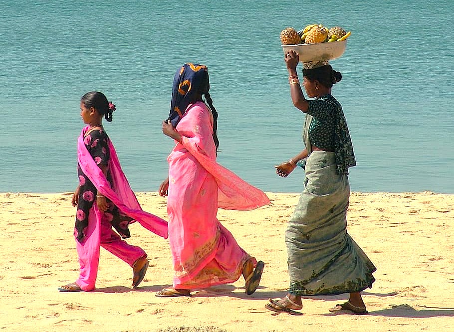 Local women in colorful sarees walk a sandy beach in Goa, India, a common coastal scene near the historic Old Goa Unesco churches, including the famous Se Cathedral.