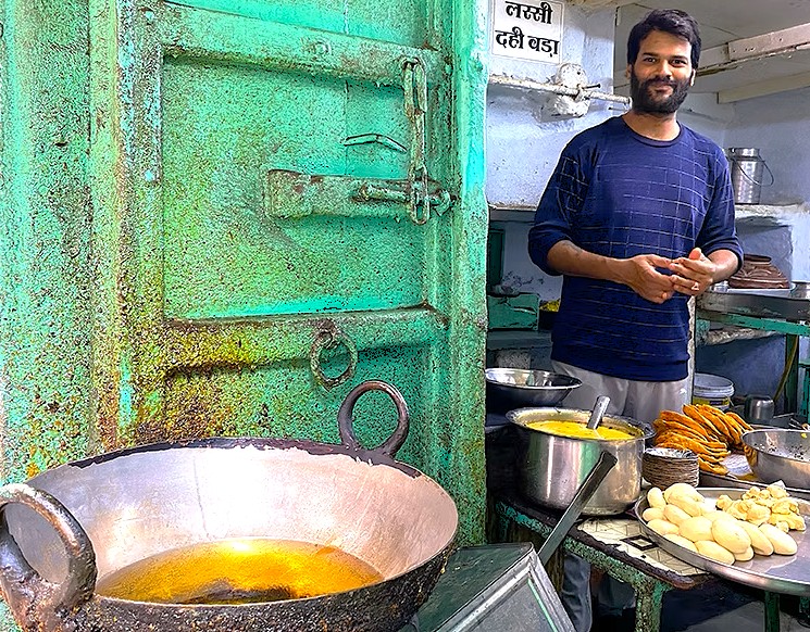 Local street food vendor prepares kachoris and lassi at a bustling Udaipur roadside stall near Taj Lake Palace in Rajasthan, India, surrounded by colorful utensils, cookware, and vibrant market ambience.