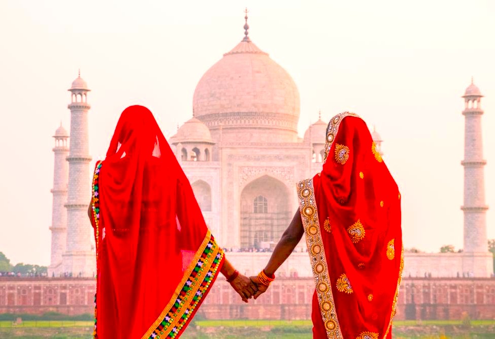 Two women in vibrant red saris hand-in-hand viewing the Taj Mahal UNESCO World Heritage Site in Agra, India, near Agra Fort and Keoladeo National Park.