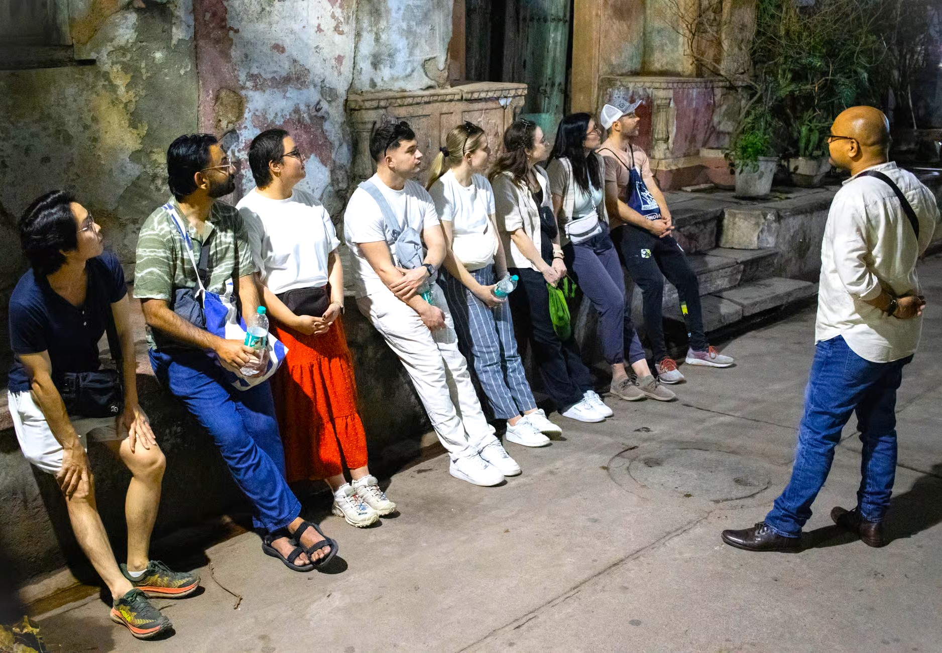 Group of ten tourists listen to a guide against a weathered Old Delhi wall during a nighttime walking tour near India Gate in India-Delhi, highlighting cultural heritage and immersive exploration. Group of ten tourists listen to a guide against a weathered Old Delhi wall during a nighttime walking tour near India Gate in India-Delhi, highlighting cultural heritage and immersive exploration.