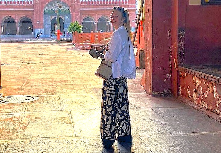 Young woman in a white shirt and patterned wide-leg pants holds a camera and book inside a pink Mughal-style courtyard in Pushkar, Rajasthan, India, with sunlight illuminating ornate arches.