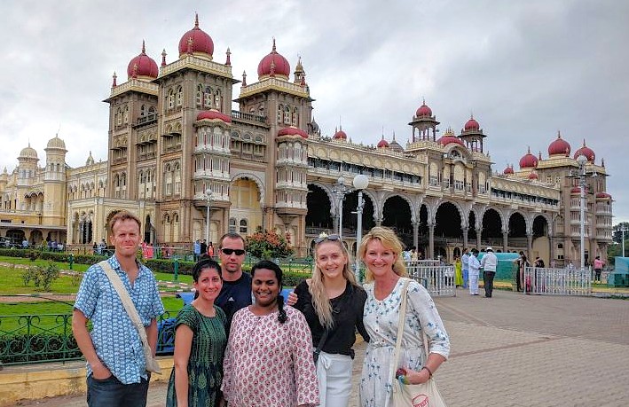 "Six smiling tourists pose before the majestic Mysore Palace in Karnataka, India, evoking the rich aroma of Mysore oils and incense and highlighting local heritage."