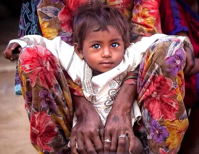 Young girl with expressive eyes sits between her mother’s knees in a rural Udaipur, Rajasthan, India setting, illustrating maternal bond, local culture, traditional attire, outdoor portrait photography capturing childhood innocence. Young girl with expressive eyes sits between her mother’s knees in a rural Udaipur, Rajasthan, India setting, illustrating maternal bond, local culture, traditional attire, outdoor portrait photography capturing childhood innocence.
