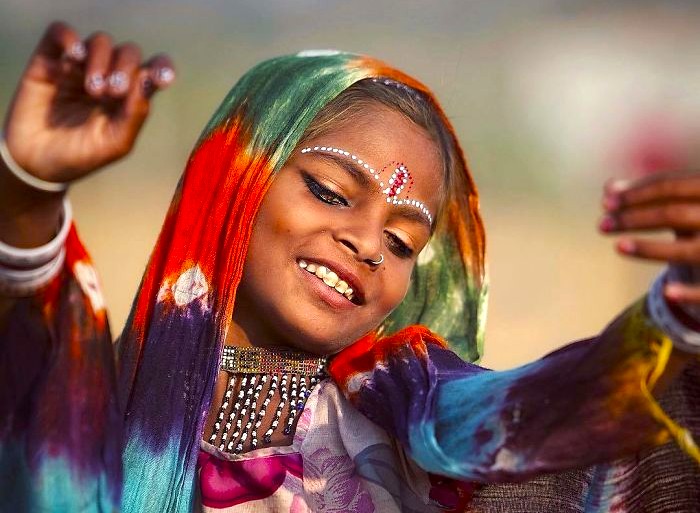 A smiling Rajasthani dancer in vibrant multicolored traditional attire, adorned with intricate face paint and jewelry, performs graceful gestures against the historic Umaid Bhawan Palace backdrop in Jodhpur, Rajasthan, India.