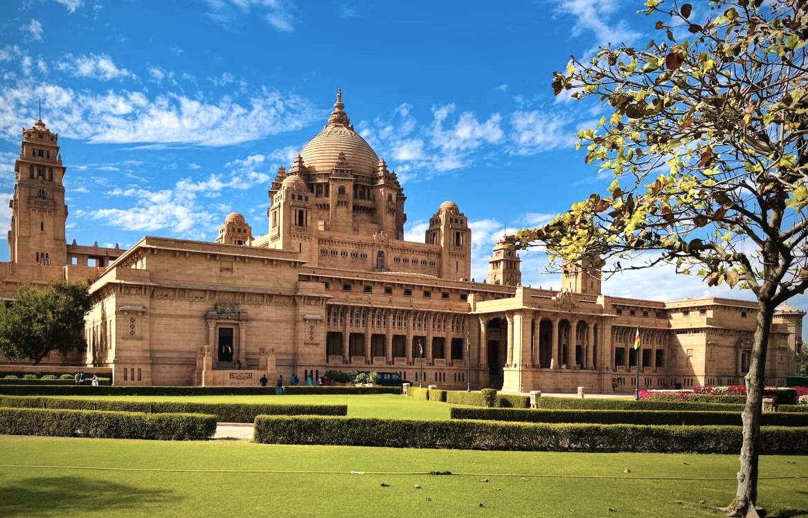 “Umaid Bhawan Palace in Jodhpur, Rajasthan, India features grand sandstone facades, majestic central dome, symmetrical towers, sunlit manicured gardens, intricate carvings, royal heritage museum, panoramic views of the city skyline.”
