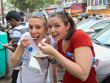 Two cheerful female tourists savor traditional Rajasthani street food near Umaid Bhawan Palace in Jodhpur, Rajasthan, India, capturing authentic, colorful local culinary culture against the backdrop of historic sandstone architecture.
