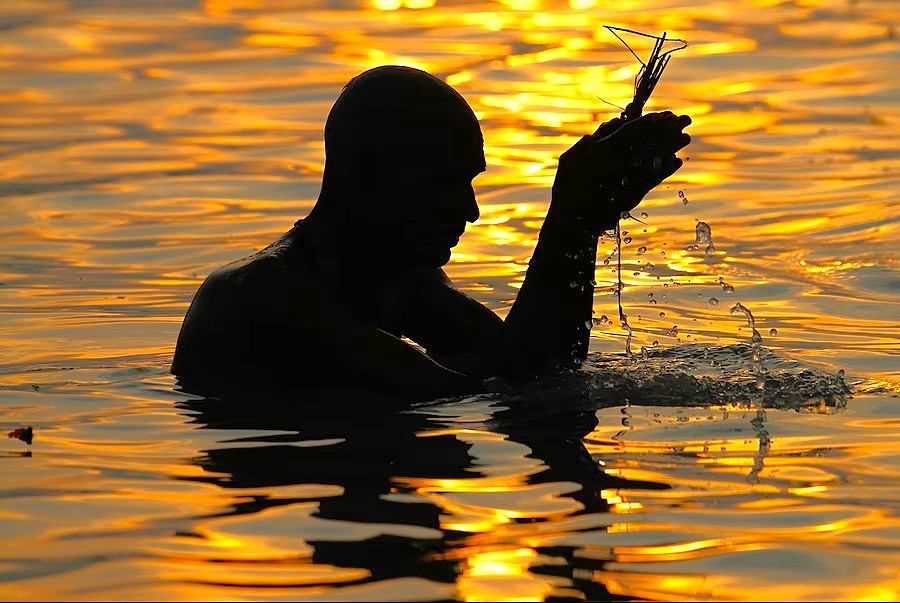 Silhouetted devotee gently lifts sacred water from the golden-hued Ganges at serene sunrise, symbolically cradling a delicate insect near Dasaswamedh Ghat in Varanasi, India, close to the historic Sarnath-Unesco site.