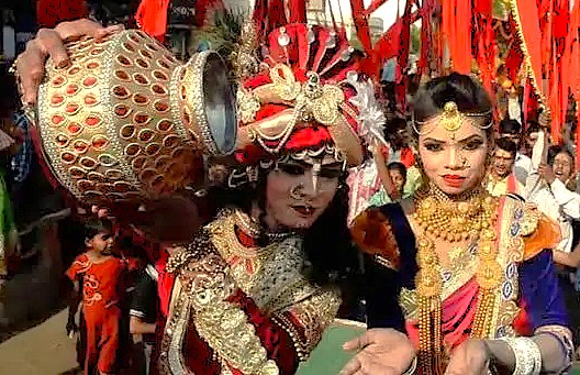 Two elaborately dressed performers in Varanasi, Uttar Pradesh, India, near Sanarth Temple Buddha, participate in a vibrant Krishna festival procession, showcasing ornate costumes, ceremonial pots, colorful decorations, and joyful crowds.