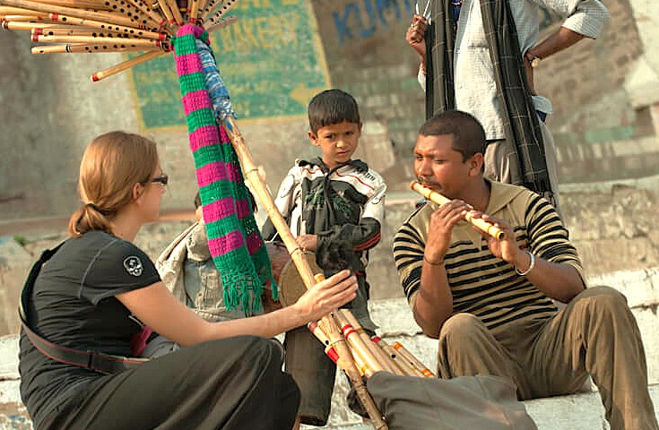 “In Varanasi, Uttar Pradesh, India, a street musician plays bamboo flute for a tourist near Sarnath Temple Buddha and Bharat Kala Bhavan Museum, showcasing vibrant cultural heritage and local artistry.”